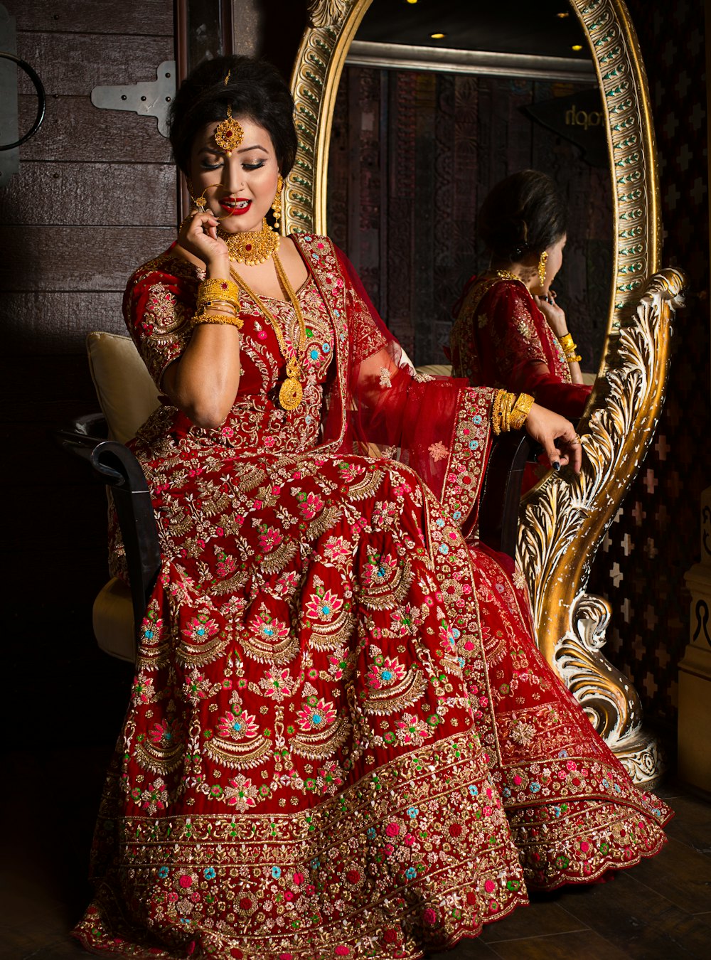 A bride in a red embroidered lehenga beside an ornate mirror
