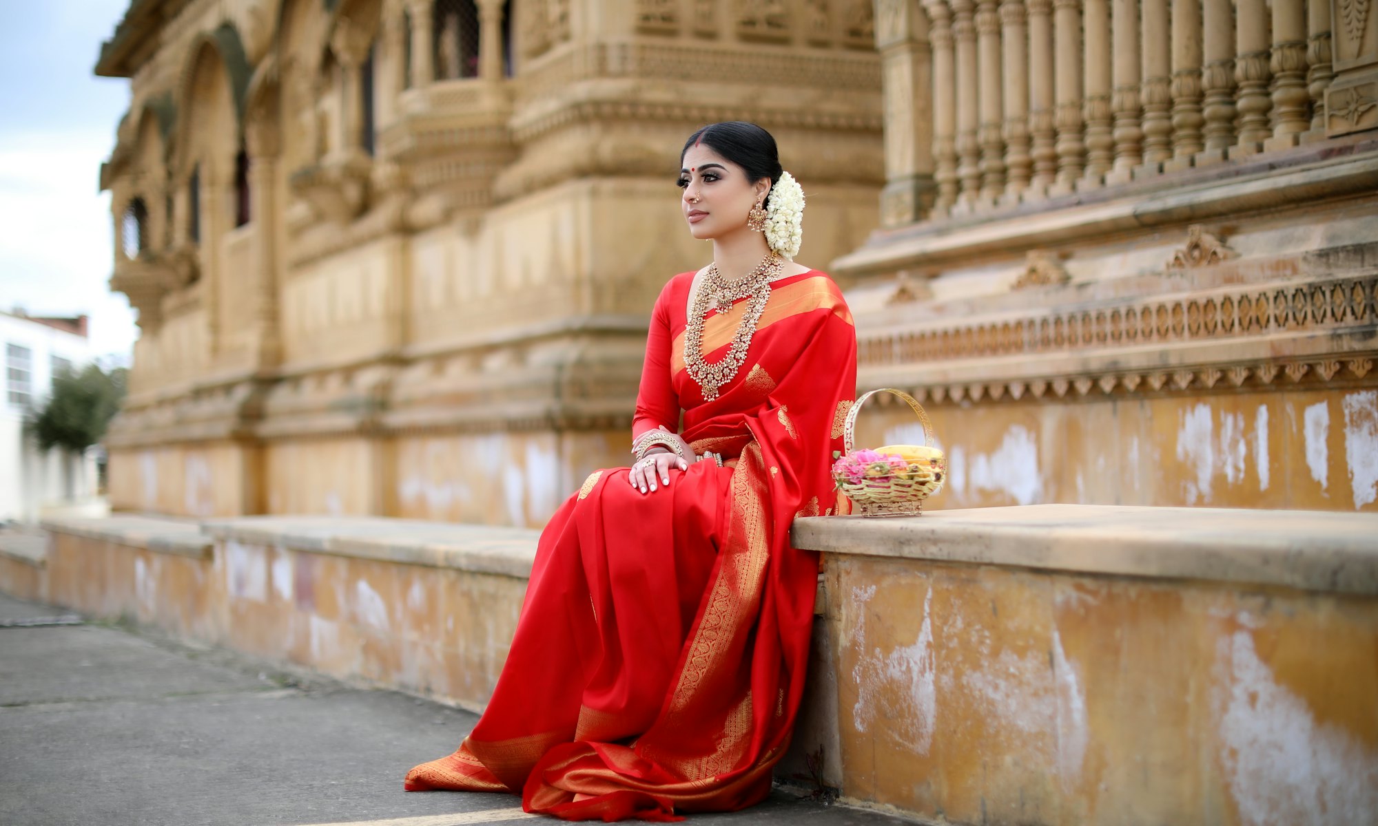 A woman in a red silk saree seated on the steps of a sandstone haveli